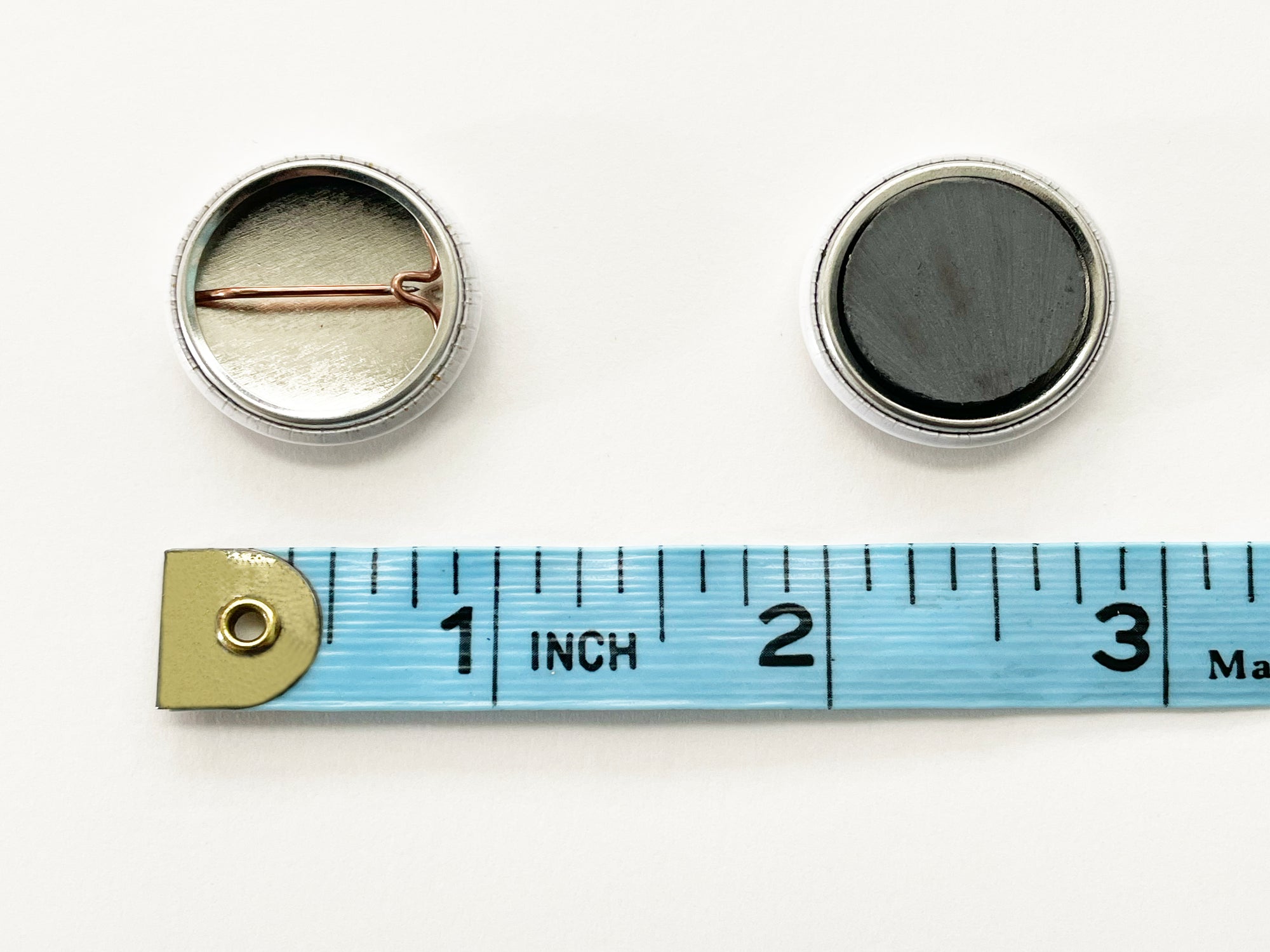 Backs of 1" pin back button and 1" magnet, shown with measuring tape for scale