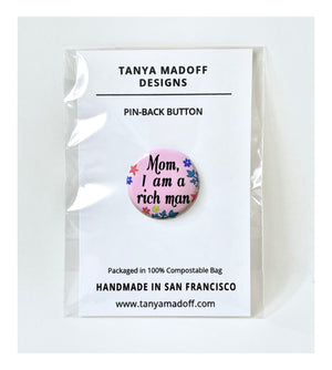 Mom, I Am A Rich Man one inch pin back button, Cher quote featured in Taylor Swift video, with black cursive lettering on pink background with flowers and leaves, packaged on recycled paper, in 100% compostable clear glassine bag